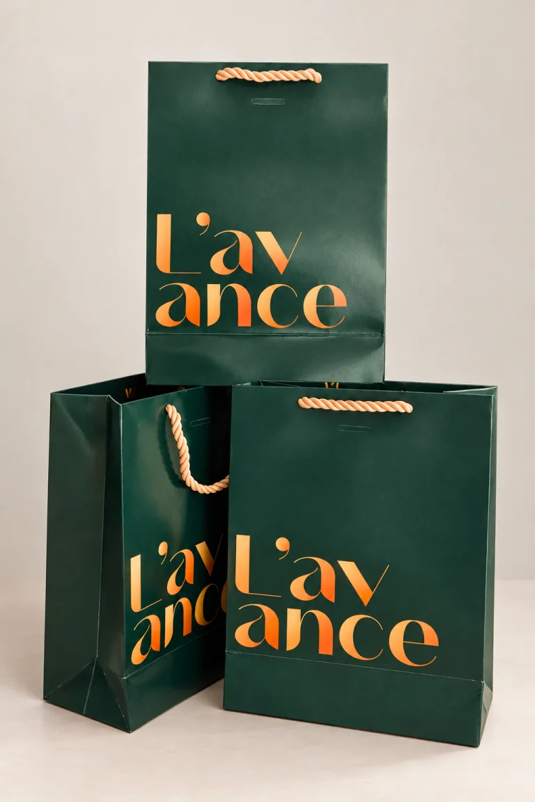 Três sacolas premium L’avance em papel verde escuro com impressão laranja e alças de cordão.